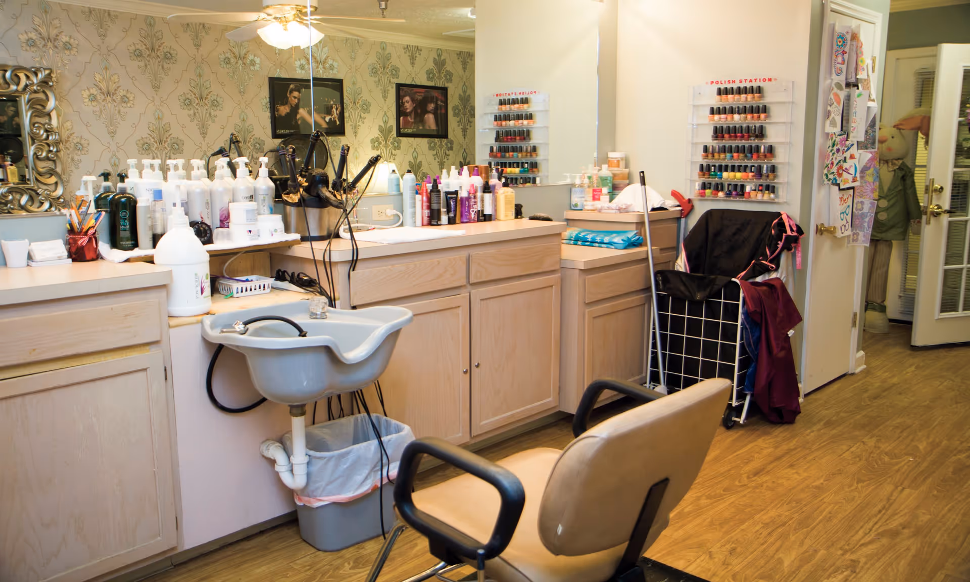Interior view of a salon area with a beige salon chair in front of a small white sink. The countertop behind the sink holds various hair care products, bottles, and styling tools. On the wall, there are racks displaying numerous nail polish bottles. The room has light wood cabinetry and a large mirror reflecting the space. The floor is wooden, and there is a cart with cleaning supplies and a mop nearby.