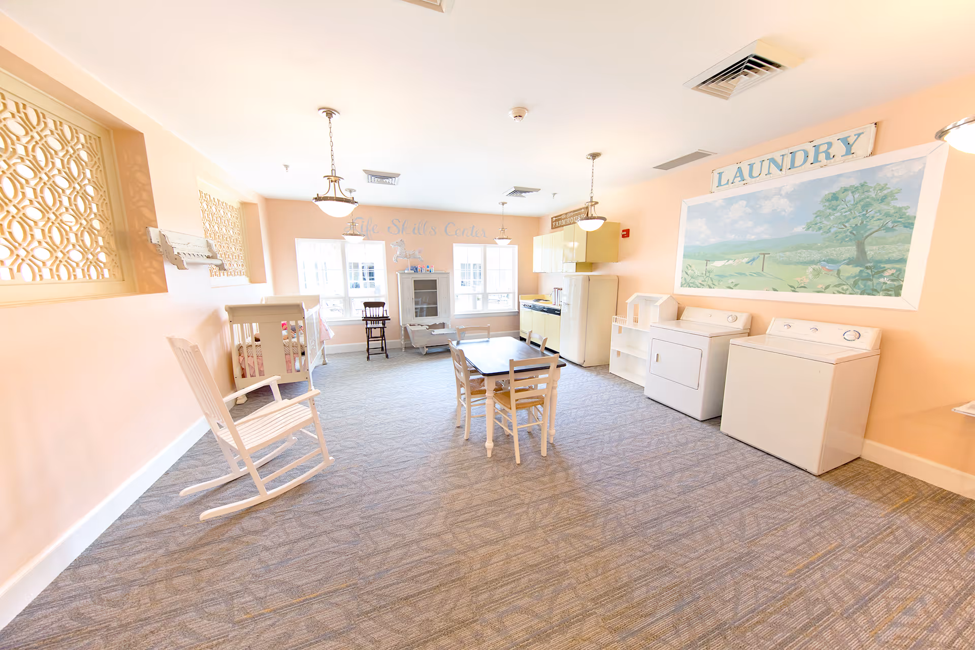 A bright, spacious room with pastel peach walls and carpeted floor, featuring a white rocking chair, a small table with four chairs, a crib, a high chair, a vintage-style refrigerator and stove, a washer and dryer, and a large mural with the word 'LAUNDRY' above it. The room is labeled as a Life Skills Center and has windows letting in natural light.