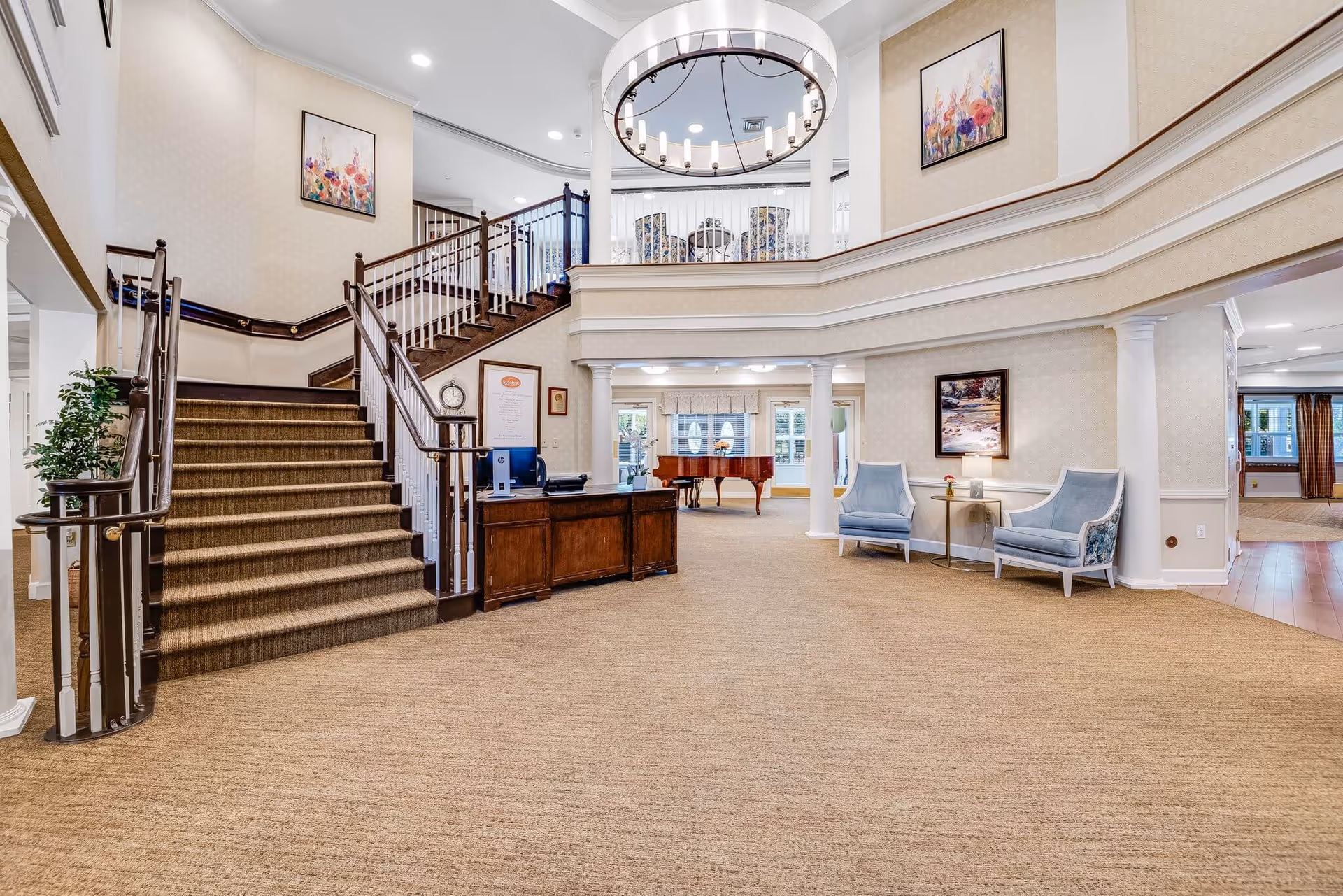 Spacious and elegant interior of a senior living facility featuring a wide carpeted area with a wooden staircase leading to an upper level. The area includes a reception desk, two blue upholstered chairs with a small table between them, framed artwork on the walls, a grand piano in the background, and a large circular chandelier hanging from the ceiling.