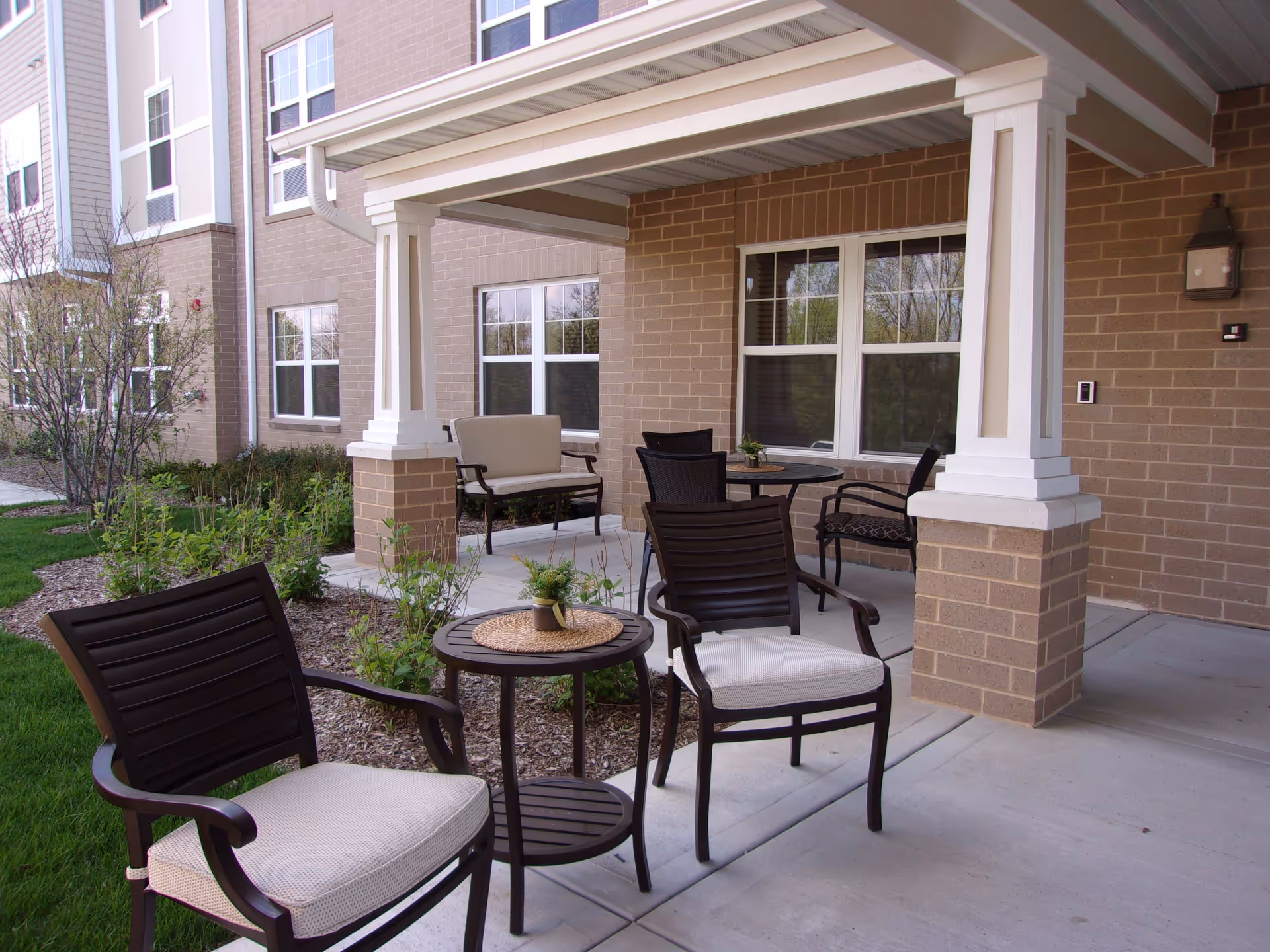 Outdoor patio area at The Whitley of Aurora featuring cushioned chairs and small tables under a covered porch attached to a brick building with windows and landscaping.