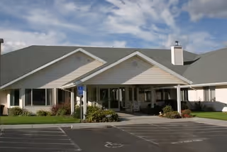 Exterior view of a single-story senior living facility building with a covered entrance, wheelchair accessible parking spaces, and a well-maintained lawn under a partly cloudy sky.