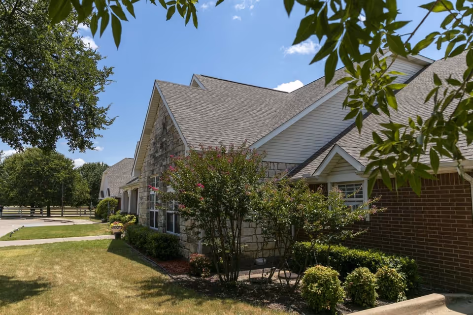 Front exterior of a single-story senior living building with stone and brick facade, landscaped shrubs and trees under a blue sky.