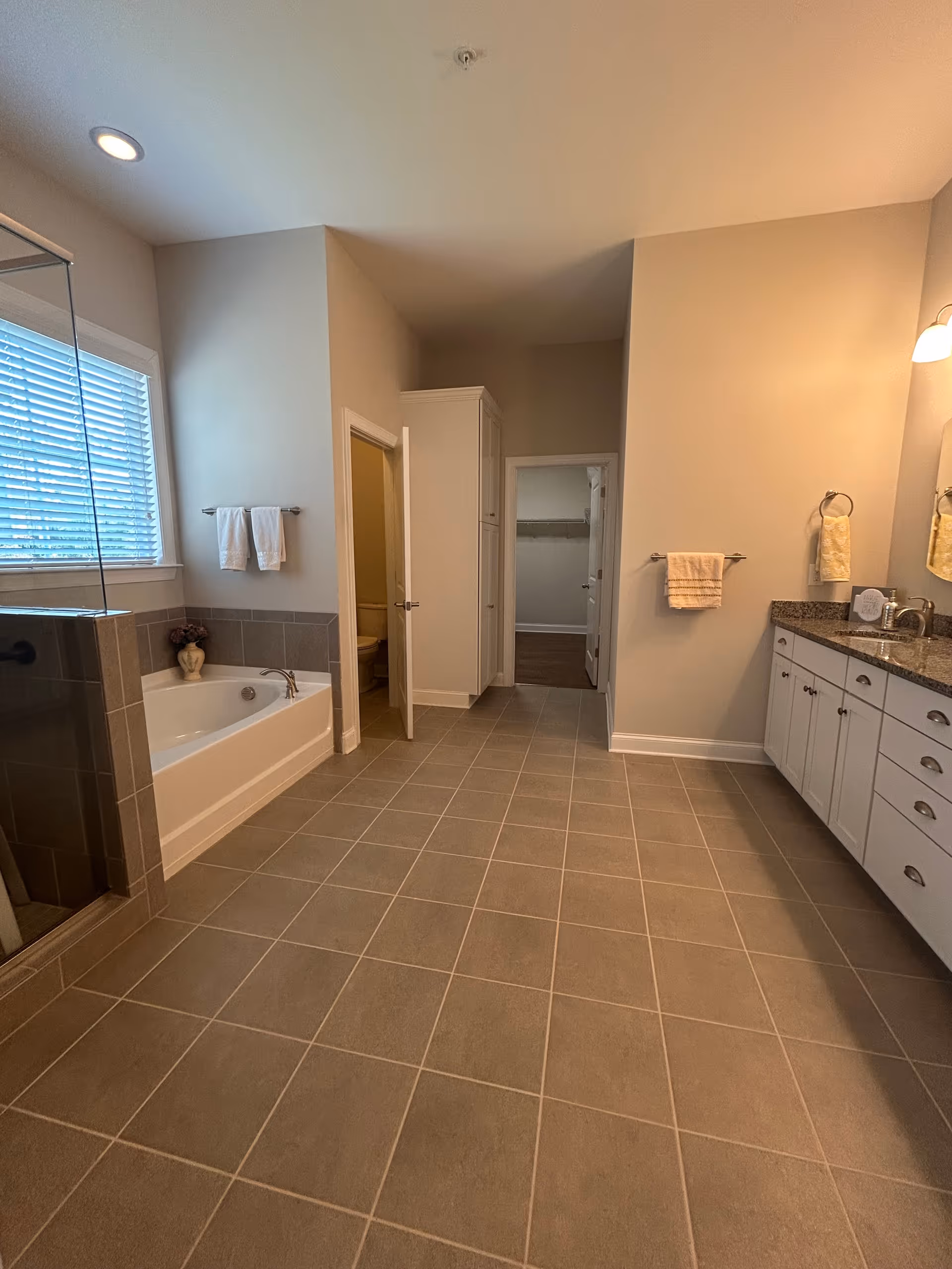 Spacious bathroom with a large bathtub on the left, a glass-enclosed shower, a double sink vanity with granite countertop on the right, beige tiled floor, and neutral-colored walls. There are towels hanging on racks and a window with blinds above the bathtub.