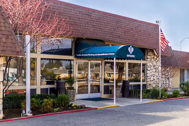 Entrance of Pacific Gardens Nursing and Rehabilitation Center with glass double doors under a green awning. The building has a stone facade and an American flag on a flagpole to the right. There are plants and shrubs near the entrance and a clear sky above.
