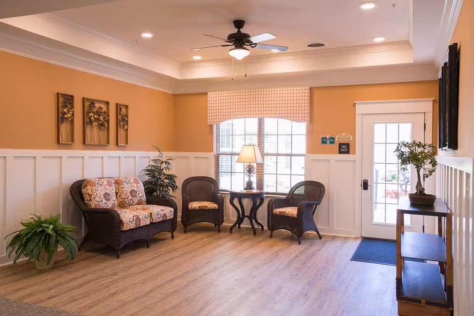 A cozy sitting area in a senior living facility with a wicker loveseat and two wicker chairs with floral cushions arranged around a small round table with a lamp. The room has light wood flooring, beige walls with white wainscoting, a ceiling fan with light, and a large window with a patterned valance. There are decorative wall art pieces, potted plants, and a door with a 'No Exit' sign.