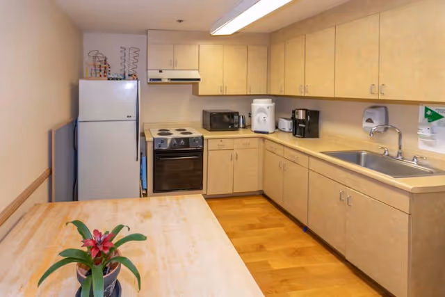 A small kitchen area with light wood cabinets and countertops, a white refrigerator, an electric stove with oven, a microwave, a coffee maker, a water dispenser, and a sink. There is a wooden table in the foreground with a potted plant on it. The floor is wooden and the walls are light-colored.