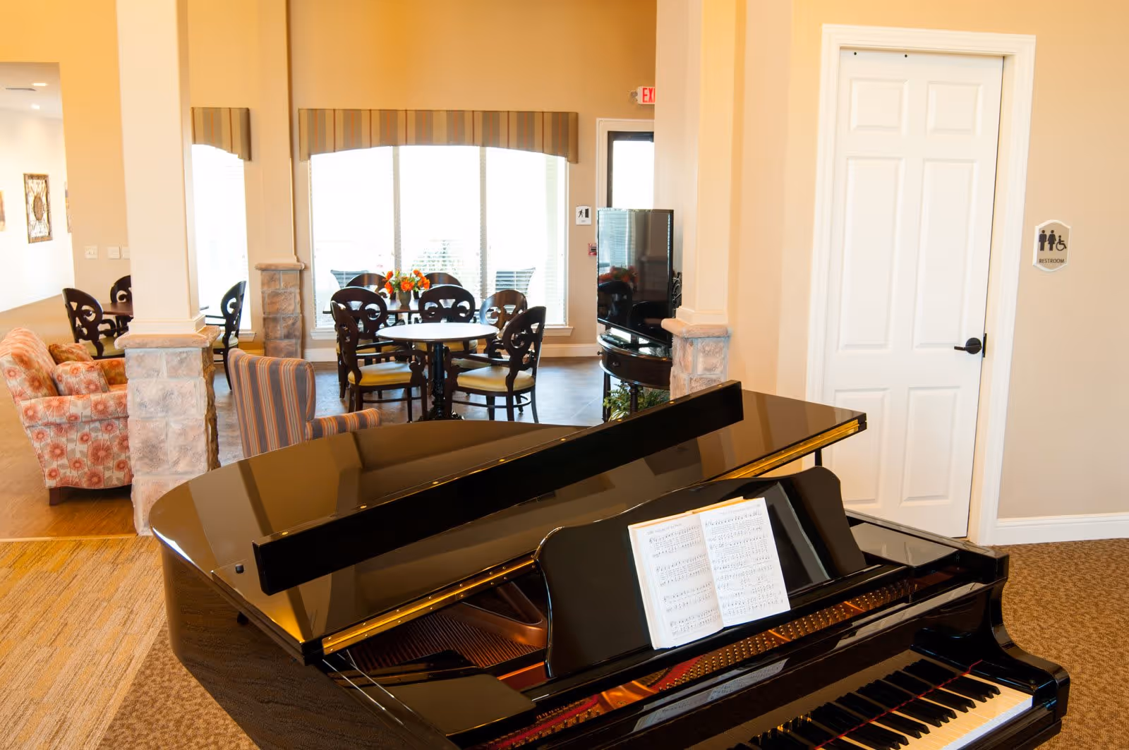 Interior view of a senior living facility lounge area featuring a black grand piano with sheet music, a seating area with patterned armchairs, a round table with chairs near large windows, and a television. The walls are painted beige and there is a door labeled as a restroom.