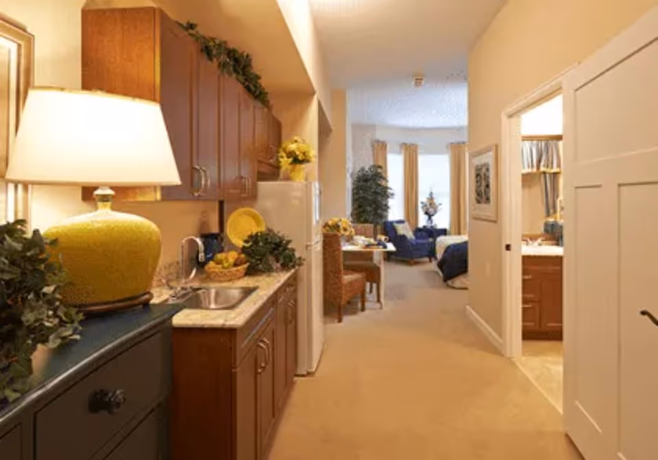 Interior view of a senior living facility apartment showing a kitchenette with wooden cabinets, a small sink, and a countertop with a basket of fruit and decorative plants. To the right, there is an open door leading to a bathroom. In the background, a dining area with chairs and a table is visible, along with a living space featuring armchairs and large windows with curtains.