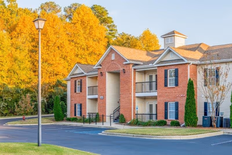 Exterior view of a two-story senior living facility building with red brick and beige siding, surrounded by trees with autumn foliage. The building has balconies, black shutters, and a small parking area in front with a streetlamp.