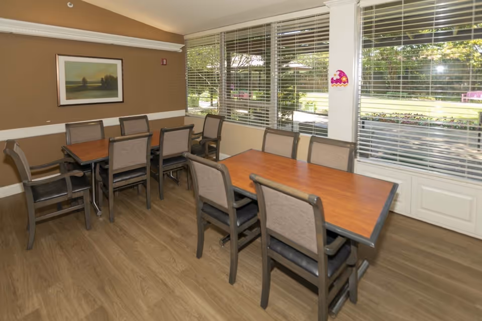 A dining room with two wooden tables and eight chairs. The room has large windows with white blinds that look out onto a green outdoor area. There is a framed landscape painting on the brown wall and wood flooring.