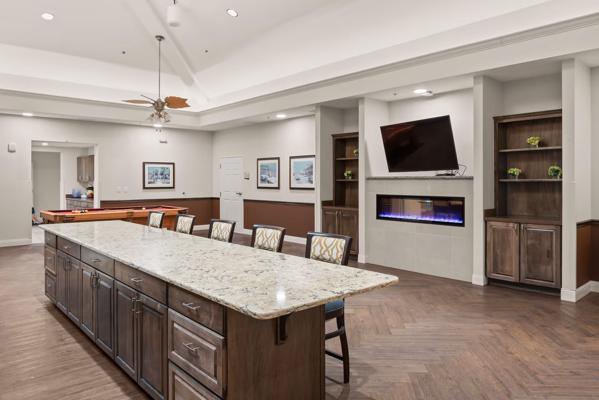A spacious senior living common area featuring a large granite countertop island with multiple chairs, a wall-mounted flat-screen TV above a modern electric fireplace, built-in wooden shelves with small plants, a ceiling fan with leaf-shaped blades, and a pool table in the background.