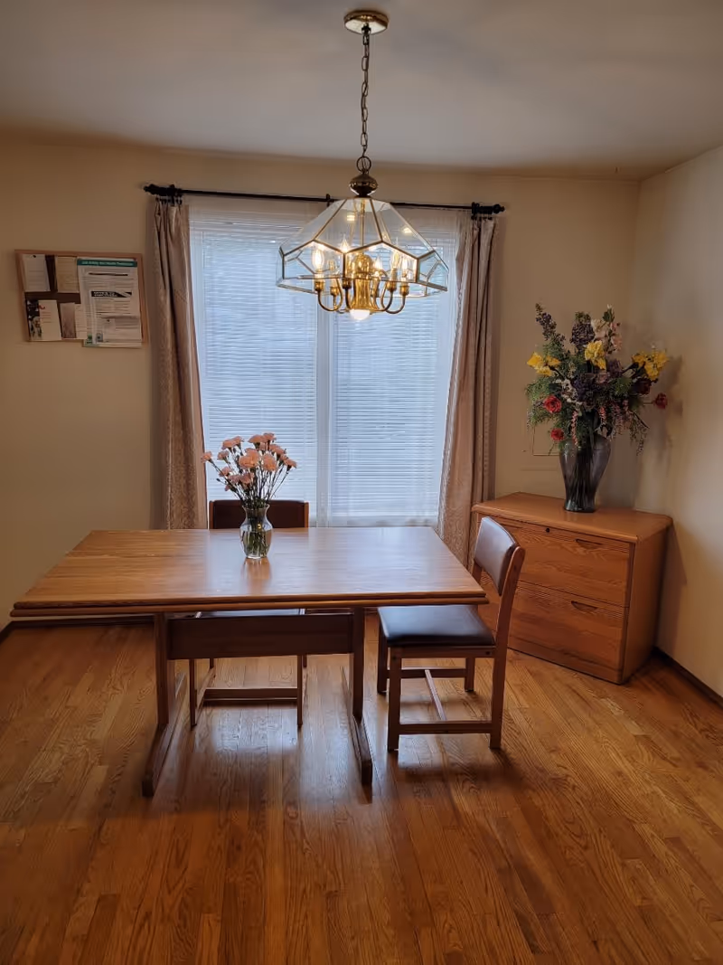 Dining room with a wooden table and chairs under a chandelier, a vase of flowers on the table and a cabinet with a floral arrangement by a window.