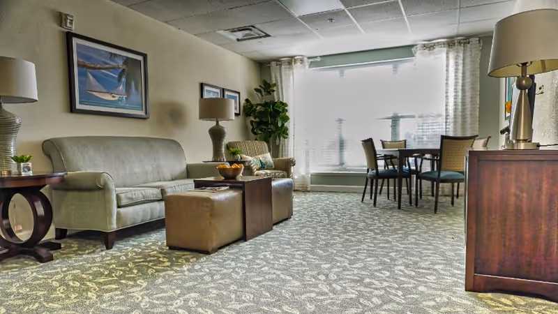 Bright communal living room with sofas, ottomans, lamps, a dining table, and a large window with curtains.