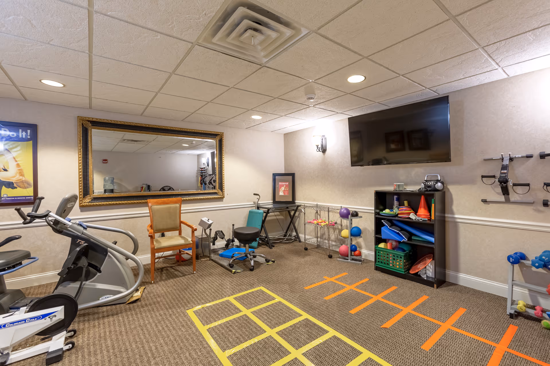 A small exercise room with various fitness equipment including a stationary bike, weights, exercise balls, and resistance bands. There is a large mirror on one wall, a TV mounted on another wall, and a chair placed near the equipment. The floor has colorful tape markings for exercise routines.