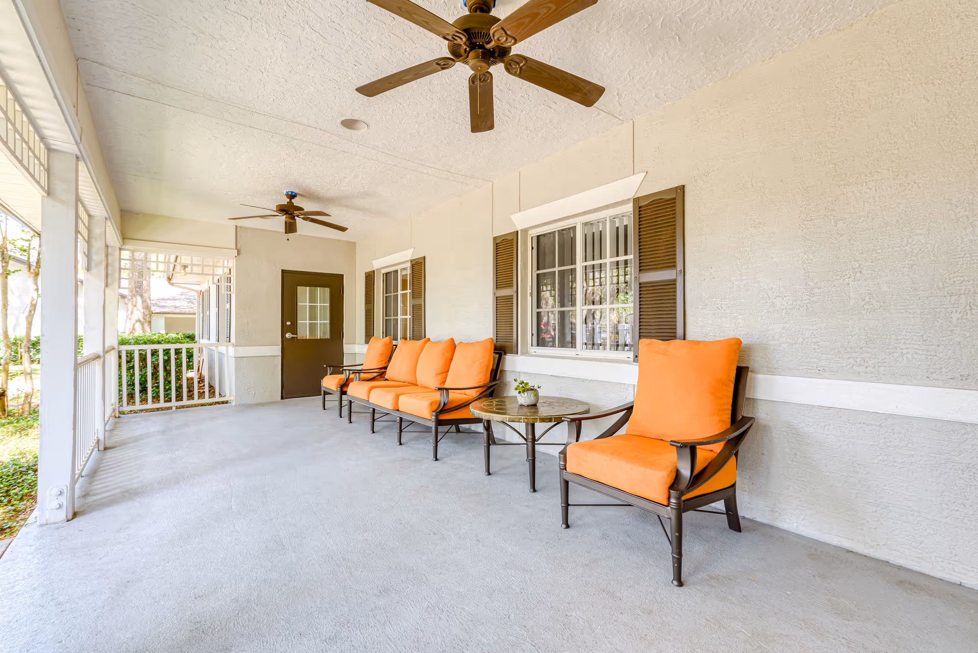 Covered porch with orange-cushioned chairs and a small table under ceiling fans overlooking a garden.
