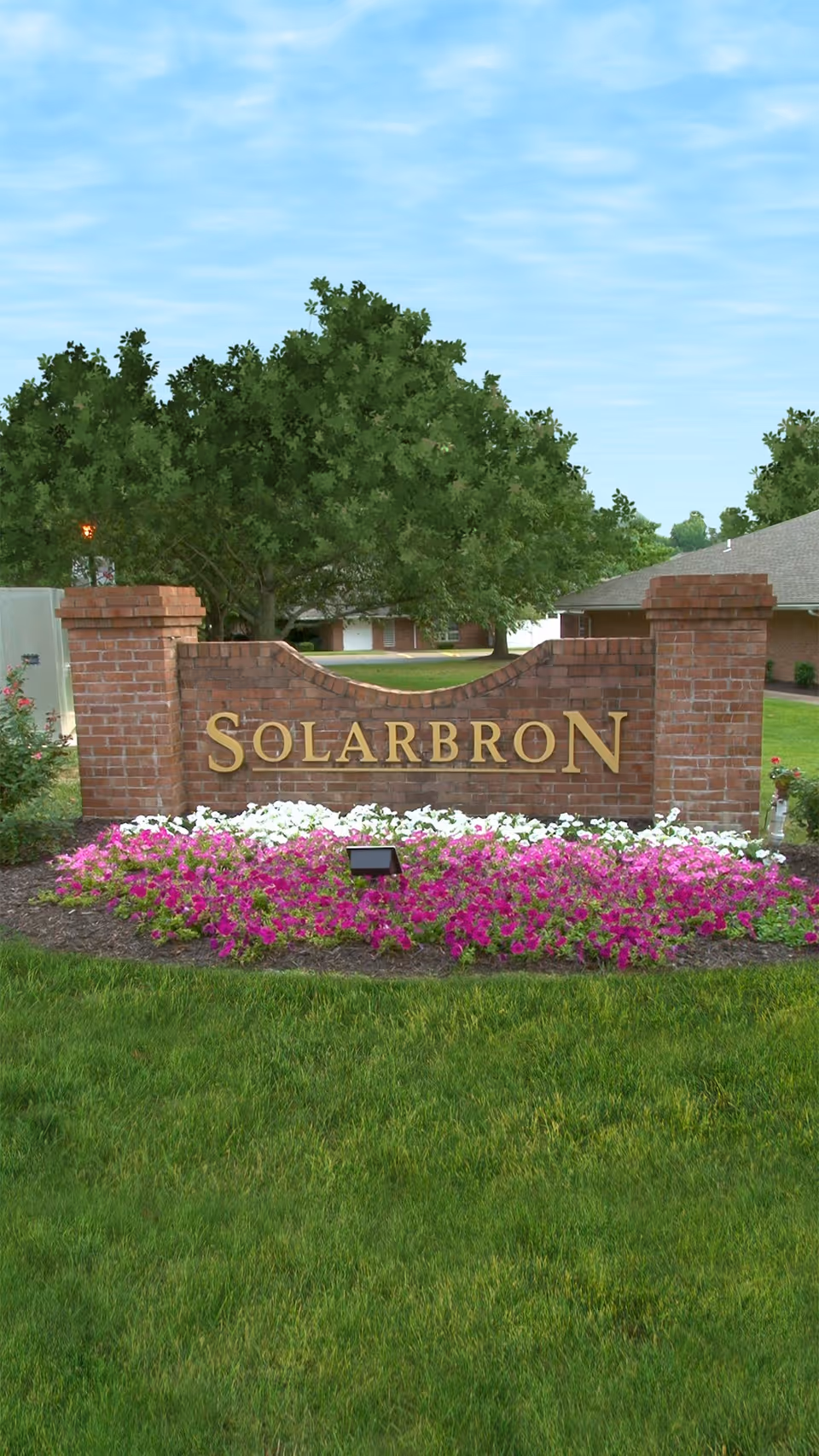 Brick entrance sign reading "SOLARBRON" set in a flowerbed with a lawn, trees, and buildings behind it.