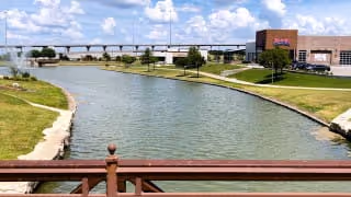 View of a calm river or canal with grassy banks on both sides, a bridge railing in the foreground, a large building on the right side, and a blue sky with scattered clouds above.