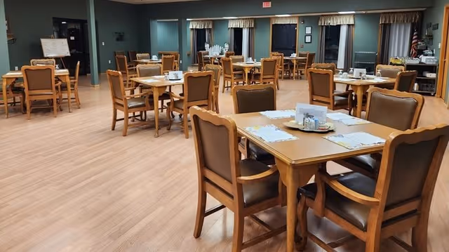 A spacious dining room with multiple wooden tables and chairs arranged neatly. Each table has placemats, napkins, and condiments. The room has large windows with curtains and a wooden floor. The walls are painted in a muted green color, and there is an American flag in the back corner.