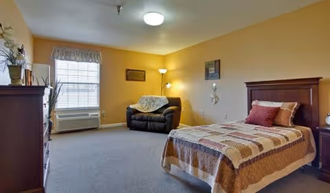 Furnished senior living bedroom with a twin bed, armchair, dresser, and a window.
