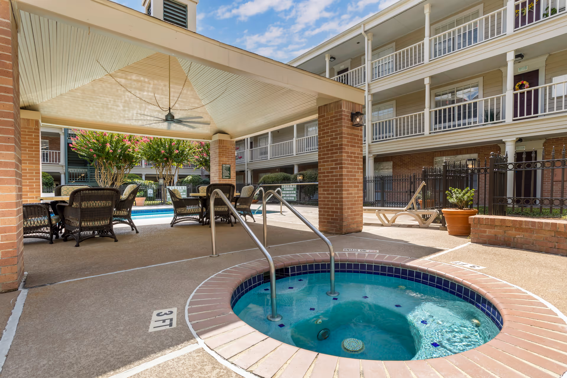Outdoor covered seating area with wicker chairs and tables next to a small round hot tub with metal handrails, surrounded by a multi-story building with balconies and potted plants under a partly cloudy sky.