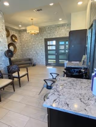 Interior view of a well-lit common area with a marble countertop in the foreground, several chairs arranged along the left wall, decorative wall hangings, a chandelier, and a set of double doors with frosted glass panels at the far end.