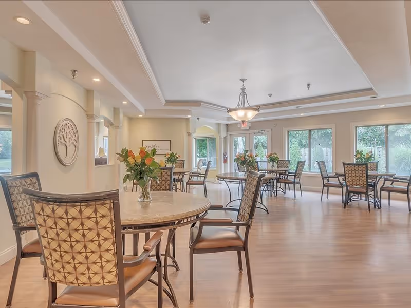 Bright, spacious dining room with round tables, patterned chairs, floral centerpieces, and large windows overlooking greenery.