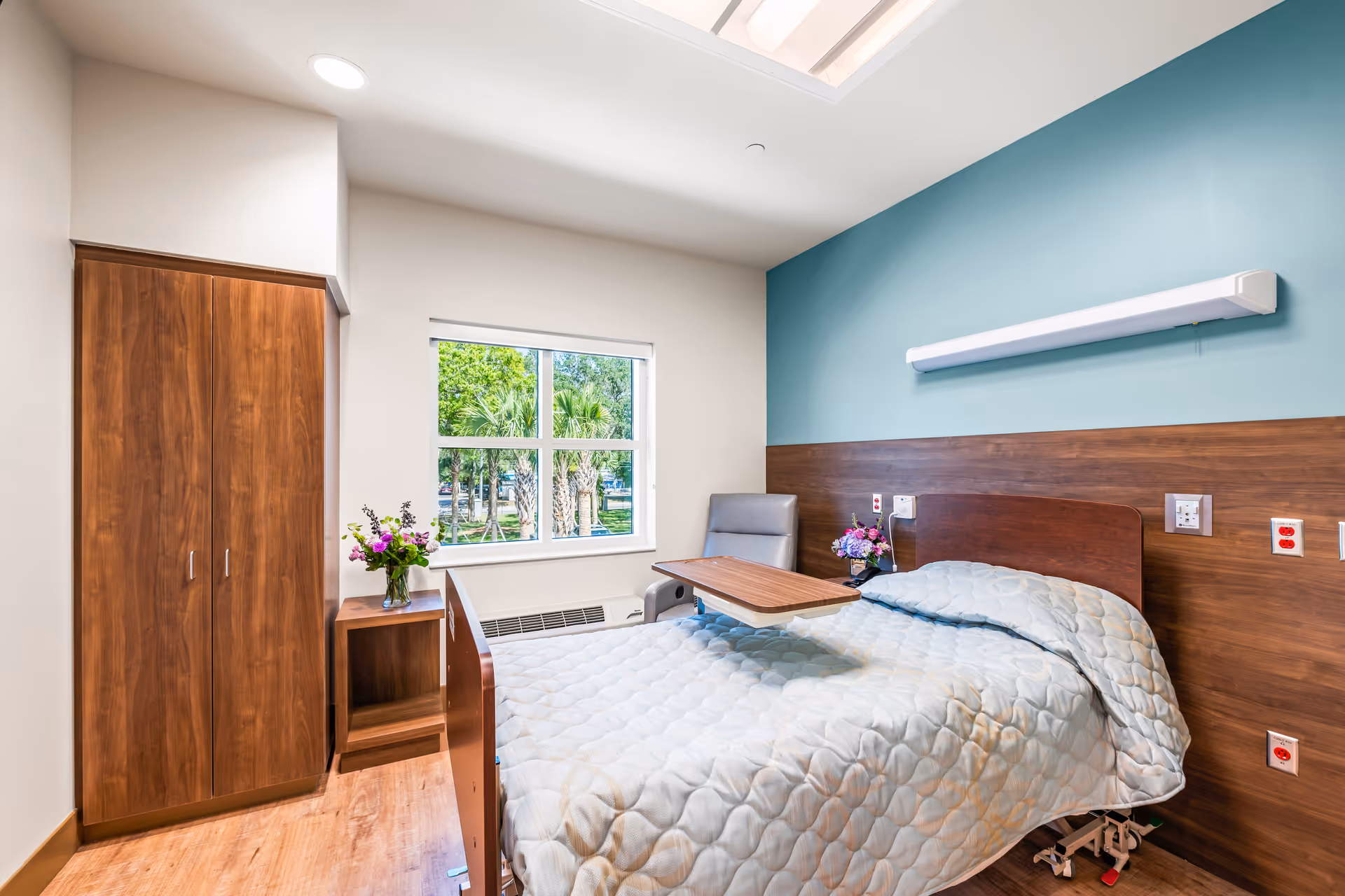 A bright and clean bedroom in a healthcare facility featuring a single hospital-style bed with a light blue quilted blanket, an adjustable overbed table, a gray armchair, a wooden nightstand with a vase of flowers, a wooden wardrobe, and a window showing green trees outside. The walls are painted white and teal with wooden paneling behind the bed.