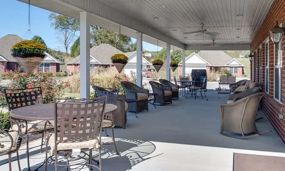 Covered outdoor patio with metal and wicker chairs, tables, and hanging planters outside a brick senior living building.