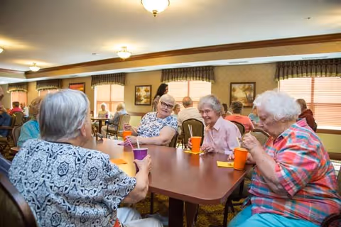 A group of elderly people sitting around a large table in a well-lit dining room, drinking from colorful cups and engaging in conversation. The room has warm lighting, framed artwork on the walls, and windows with blinds and valances.