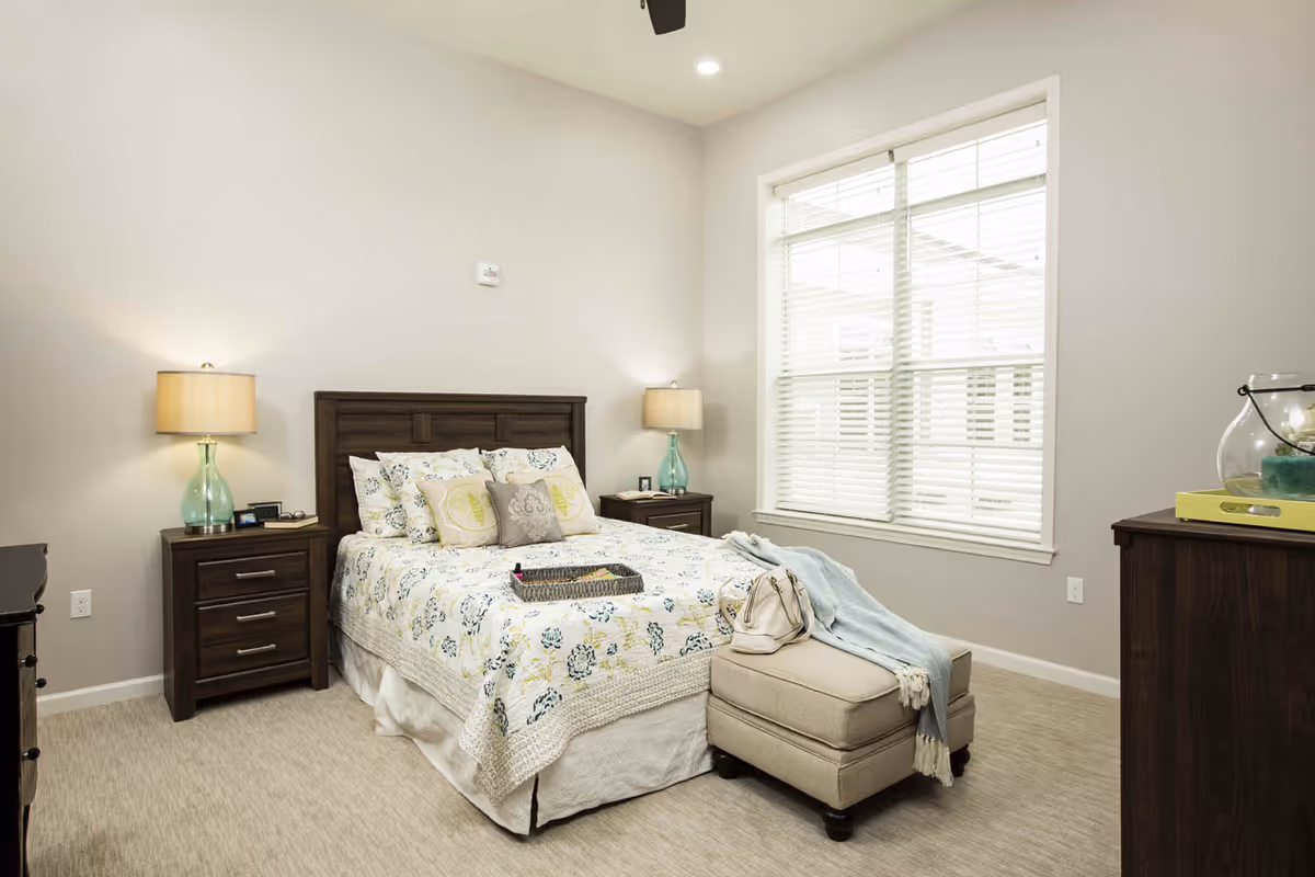 Bright, neutral bedroom with a bed framed by dark wood nightstands and lamps, a window with blinds, and an ottoman at the foot of the bed.