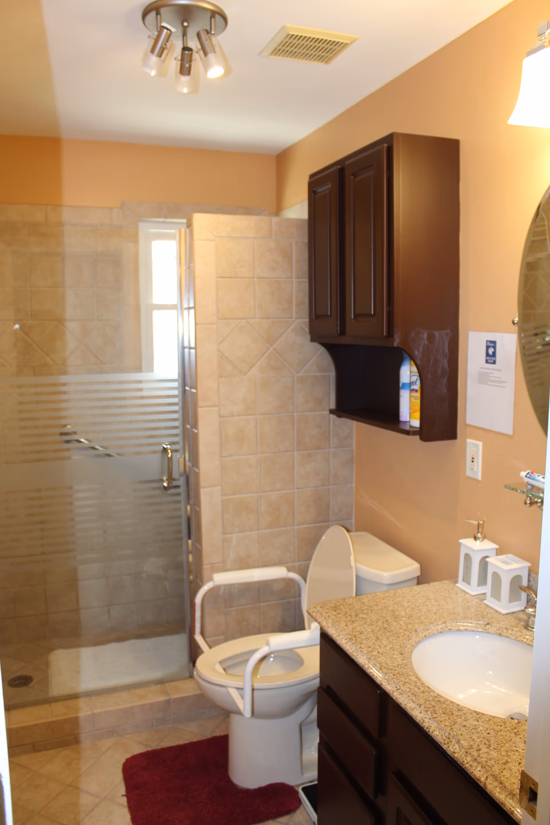A bathroom with beige tiled walls and floor, featuring a walk-in shower with a glass door, a toilet with white safety rails, a dark wood cabinet above the toilet, a granite countertop with a sink, and a round mirror on the wall. There is a red bath mat on the floor and a ceiling light fixture with multiple bulbs.