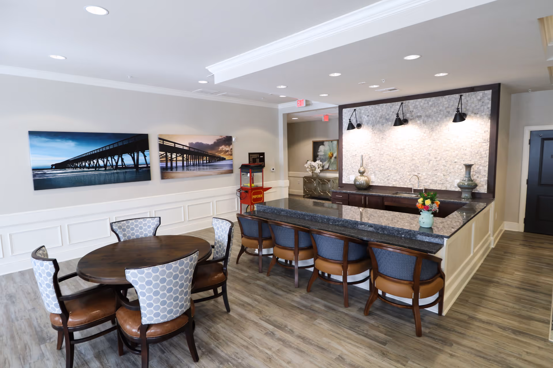 A modern dining area in an assisted living facility featuring a round wooden table with six upholstered chairs. Adjacent to the table is a granite countertop bar with five cushioned bar stools. The wall behind the bar has a textured tile backsplash with three black wall-mounted lights. Two large landscape photographs of a pier over water are hung on the wall. The floor is wood-style laminate, and there is a popcorn machine in the corner near a hallway entrance.