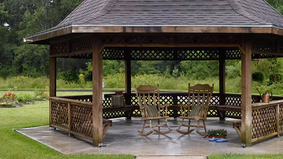 A wooden gazebo with a shingled roof situated on a concrete slab in a grassy outdoor area. Inside the gazebo are two wooden rocking chairs with cushions, a small table, potted plants, and a watering can. The background shows green trees and shrubs.