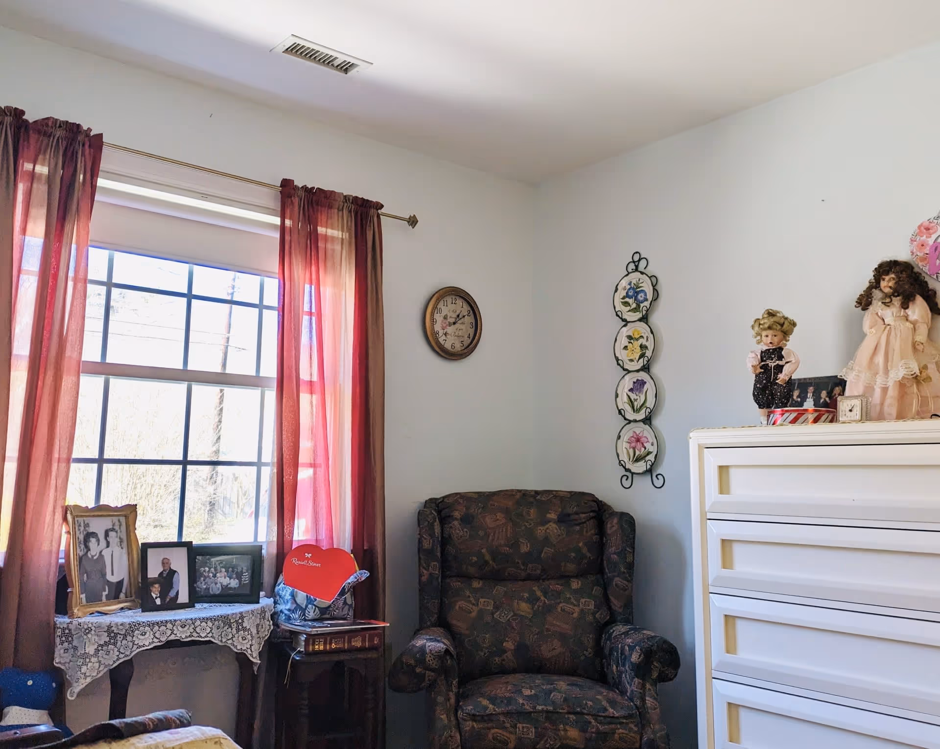 A cozy room with a window covered by sheer red curtains, a small table with framed photos and a lace tablecloth, a patterned armchair, a white dresser with decorative dolls on top, and a wall clock and floral wall decor on the walls.