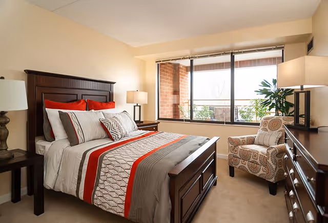 A well-lit bedroom with a large wooden bed featuring a red, gray, and white patterned comforter and multiple pillows. There are two bedside tables with lamps on either side of the bed. A patterned armchair is placed near a large window that shows an outdoor balcony with plants. A wooden dresser with a lamp and decorative items is also visible.