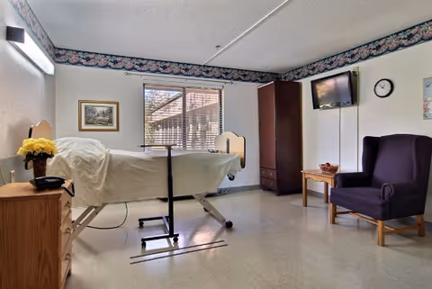 A healthcare facility room with a hospital bed covered in white sheets, a wooden nightstand with a vase of yellow flowers and a telephone, a window with blinds, a wall-mounted TV, a clock, a dark purple armchair, a small wooden table with a bowl, and a tall wooden cabinet. The room has a floral wallpaper border near the ceiling.
