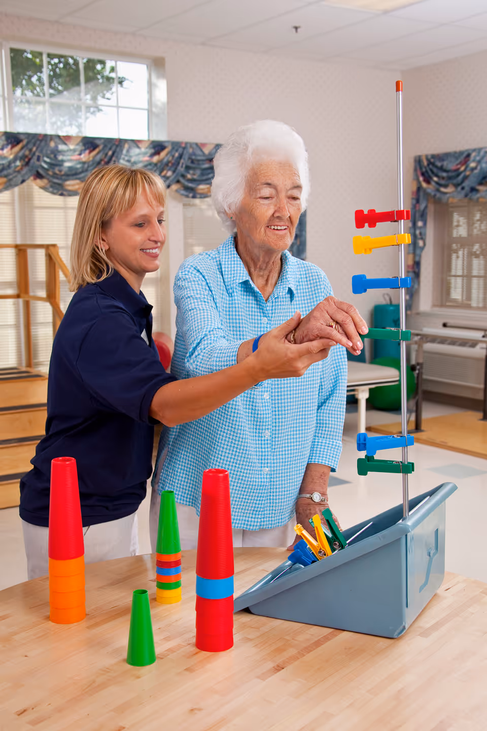 A senior woman in a blue checkered shirt is assisted by a caregiver in a navy blue shirt as they engage in a therapeutic activity with colorful plastic cups and a vertical peg board in a bright room with windows and exercise equipment.