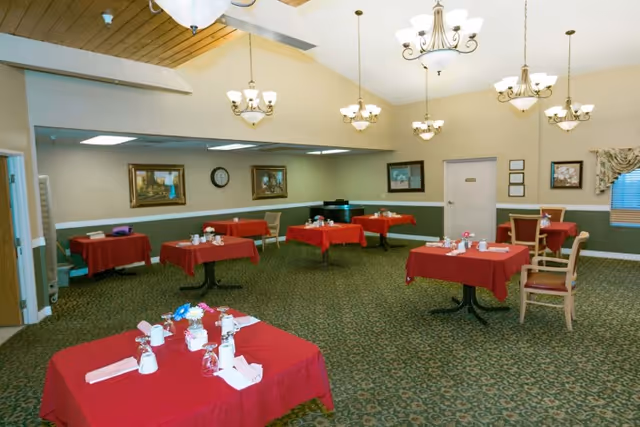 A dining room with multiple tables covered in red tablecloths, each set with white napkins, cups, and silverware. The room has green patterned carpet, beige walls with a green lower half, several framed paintings, a clock, and multiple chandeliers hanging from the ceiling. There are chairs around the tables and a piano against the far wall.