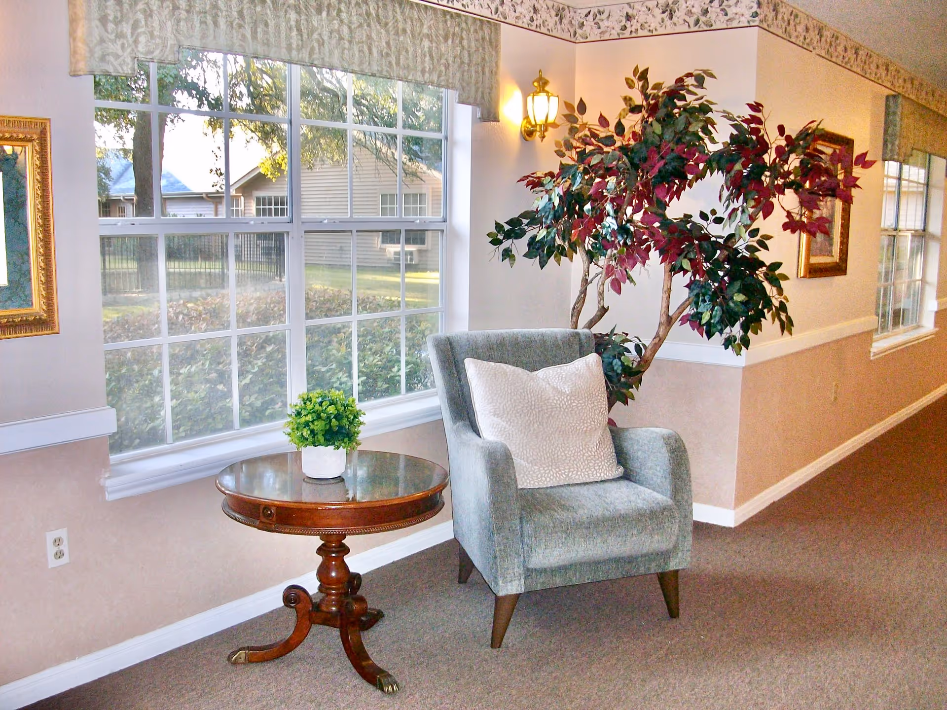 Cozy seating area with an upholstered armchair, round side table holding a potted plant, and a large window looking outside.