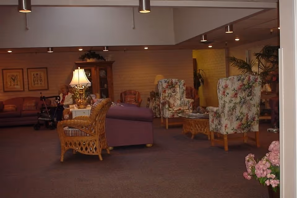 A communal living room with floral upholstered armchairs, wicker seating, a lamp, and couches in a senior living facility.
