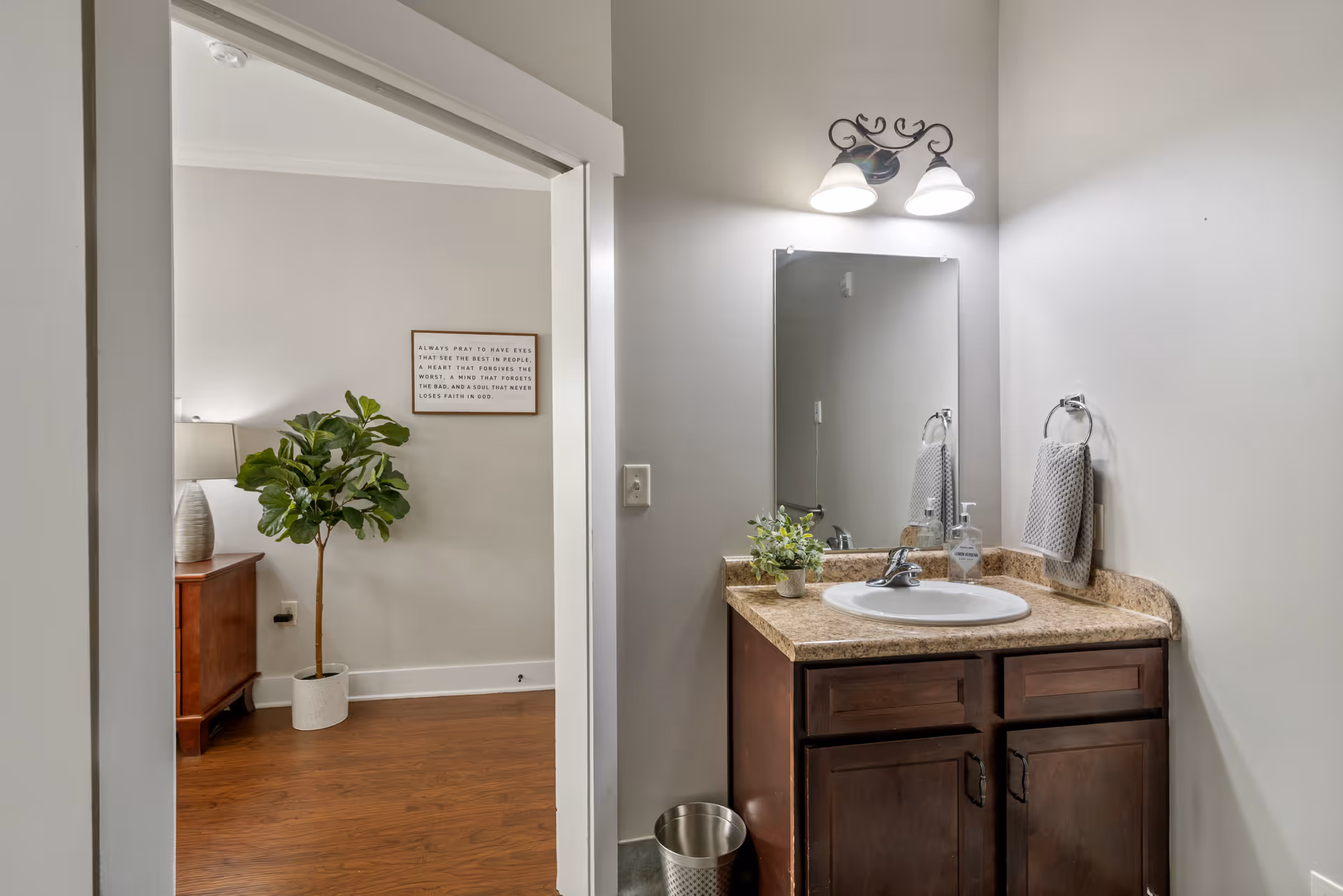 Interior view of a bathroom vanity with a sink, granite countertop, dark wood cabinets, a mirror, and a wall-mounted light fixture above. A gray hand towel hangs on a ring to the right of the sink, and a small potted plant and soap dispenser are on the countertop. To the left, an open doorway leads to a room with wooden flooring, a potted plant, a wooden dresser with a lamp, and a framed inspirational quote on the wall.