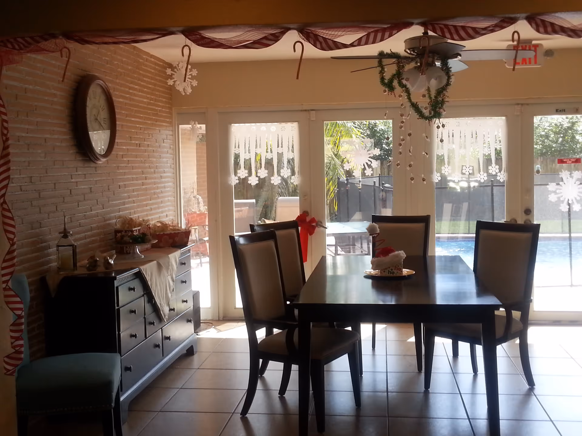 Dining area with a dark wooden table and six chairs, decorated with Christmas-themed items including hanging candy canes, snowflakes on glass doors, and a wreath on the ceiling fan. A sideboard with decorative items and a large clock on a brick wall are visible. Outside the glass doors, there is a pool and patio furniture.