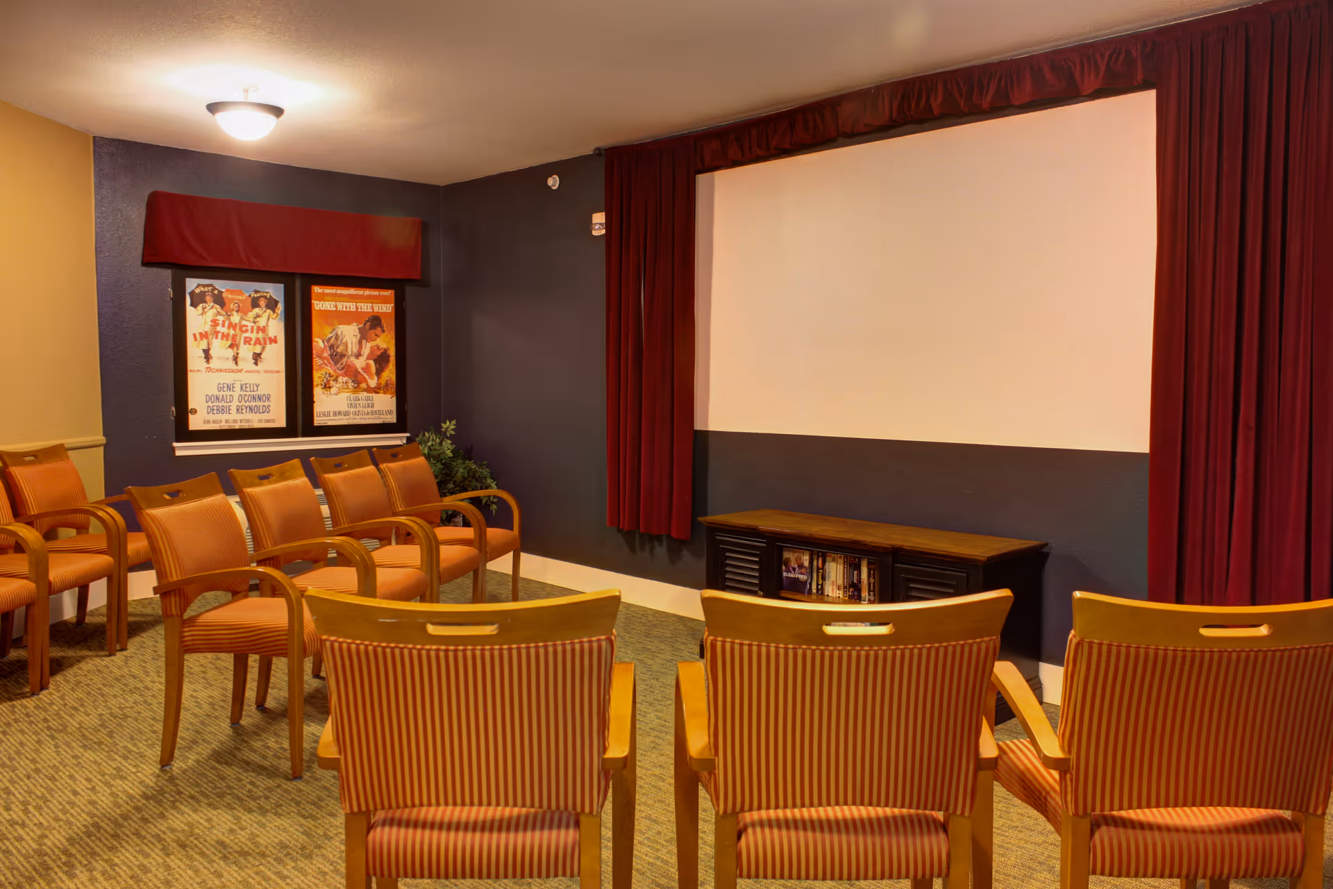 A small movie screening room with rows of wooden chairs with orange striped cushions facing a large blank projector screen framed by red curtains. Two vintage movie posters are displayed on the dark blue wall to the left, and a small cabinet with DVDs is beneath the screen.