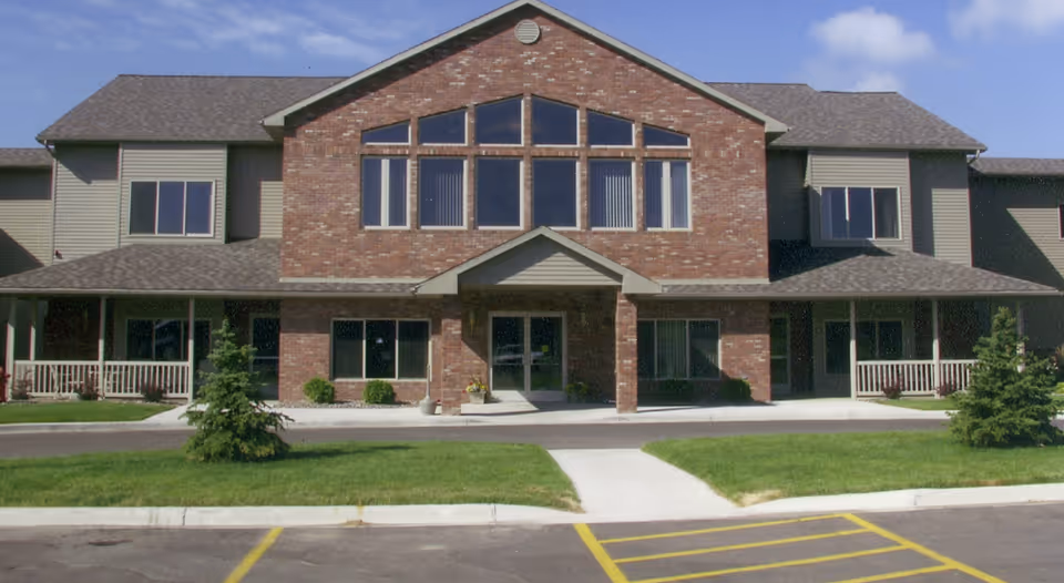Front exterior of a two-story brick and siding senior living building with a covered entrance, large windows, and a parking area.