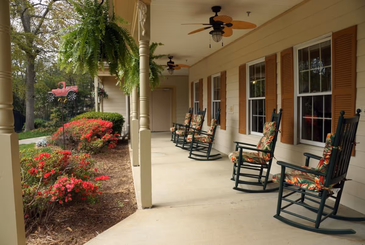 A covered outdoor porch area with several dark green rocking chairs featuring floral cushions. The porch has ceiling fans with light fixtures and is adjacent to a garden with red flowering bushes and a decorative red truck garden ornament. The building exterior is beige with brown window shutters.