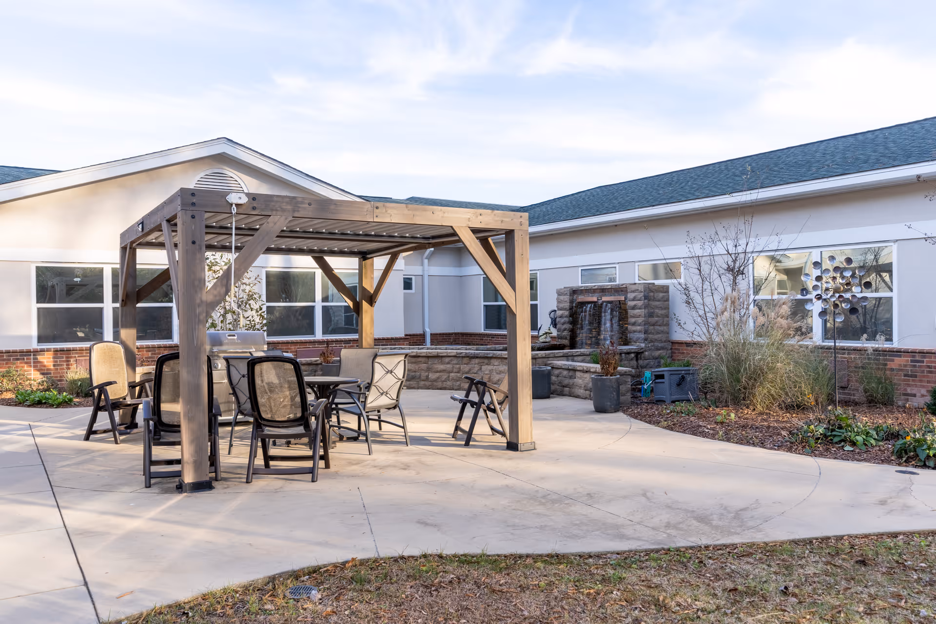 Outdoor patio area at The Center For Advanced Rehab at Parkside featuring a wooden pergola with a table and several chairs underneath. Surrounding the patio are garden beds with plants and a stone water feature against the building wall. The building has white walls with brick accents and multiple windows.