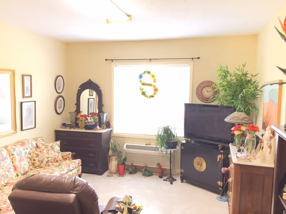A cozy living room with a floral patterned sofa, a brown recliner chair, a dark wooden dresser with a mirror, various framed pictures on the walls, a flat-screen TV on a black cabinet, several potted plants, and decorative items including flowers and figurines. A large window with a floral wreath lets in bright natural light.