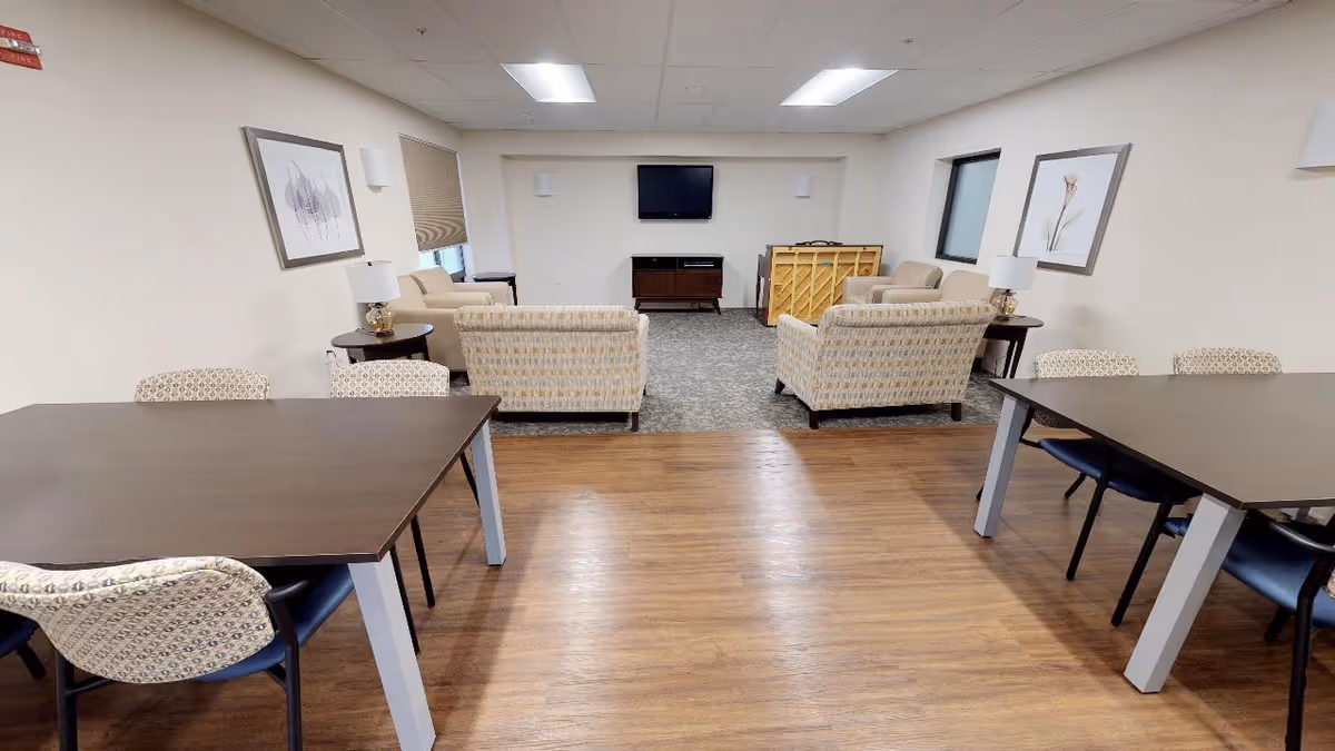 A senior living community common area with two dark wooden tables and patterned chairs in the foreground, and a seating area with beige patterned armchairs and a sofa facing a wall-mounted TV in the background. The room has light-colored walls, framed artwork, and wood flooring transitioning to carpet near the seating area.