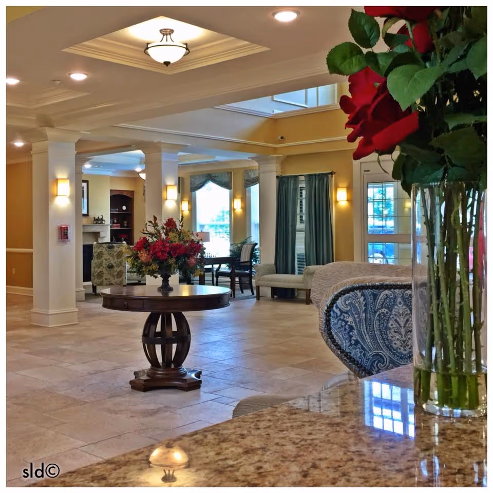 Interior view of a senior living facility lounge area with a round wooden table holding a floral arrangement in the center. The room features tiled floors, white columns, yellow walls, and multiple seating areas with upholstered chairs and sofas. Large windows with curtains allow natural light to enter, and ceiling lights illuminate the space. A vase with red roses is visible in the foreground on a granite countertop.