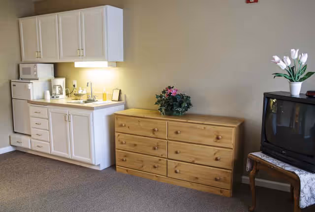 A small kitchenette area with white cabinets, a microwave, coffee maker, and sink on the left side. Next to it is a wooden dresser with six drawers and a green potted plant on top. On the right side, there is an old-style CRT television on a wooden stand with a white lace cloth and a white vase with white flowers on top.